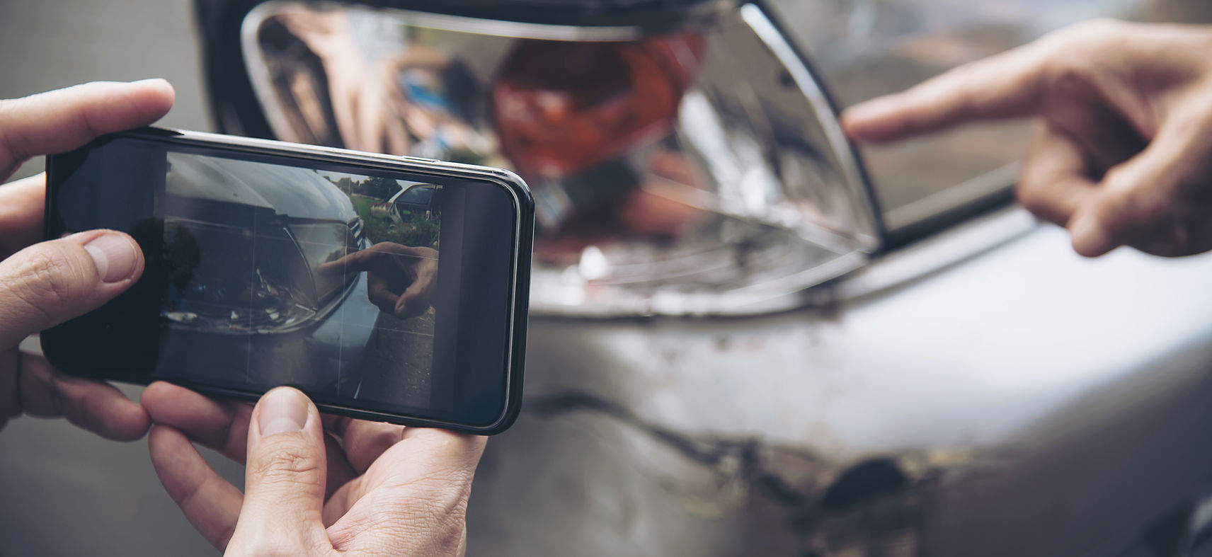 Handy macht Foto von Schaden, Hand zeigt auf beschädigtes Fahrzeug
